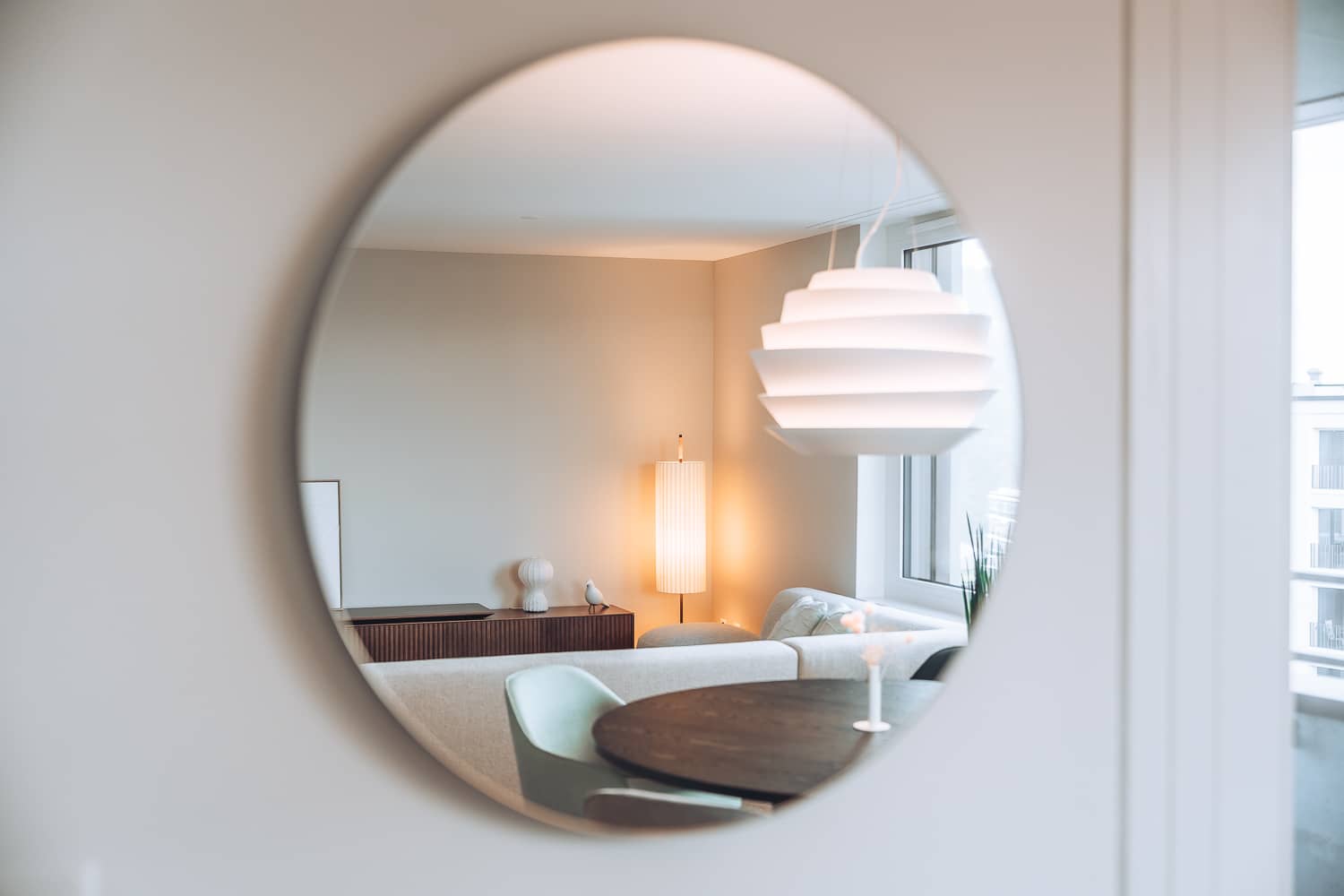 In einem runden Spiegel an der Wand spiegelt sich ein Wohnzimmer mit einer leuchtenden Stehlampe
